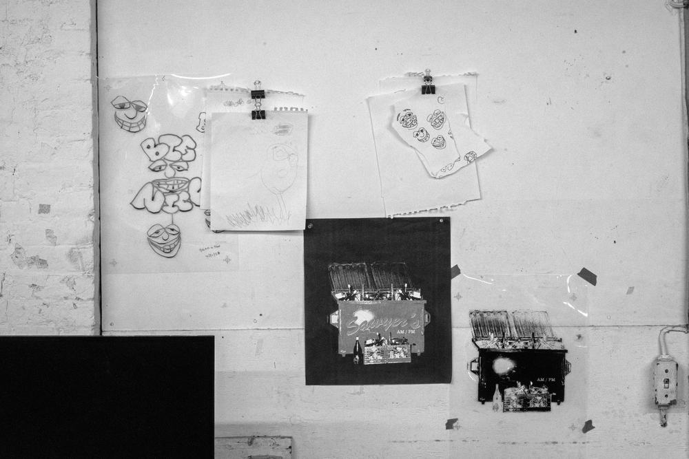 Pencil sketches and printed proofs clipped to the shop wall, including a hand-drawn dumpster illustration printed in silver on black.