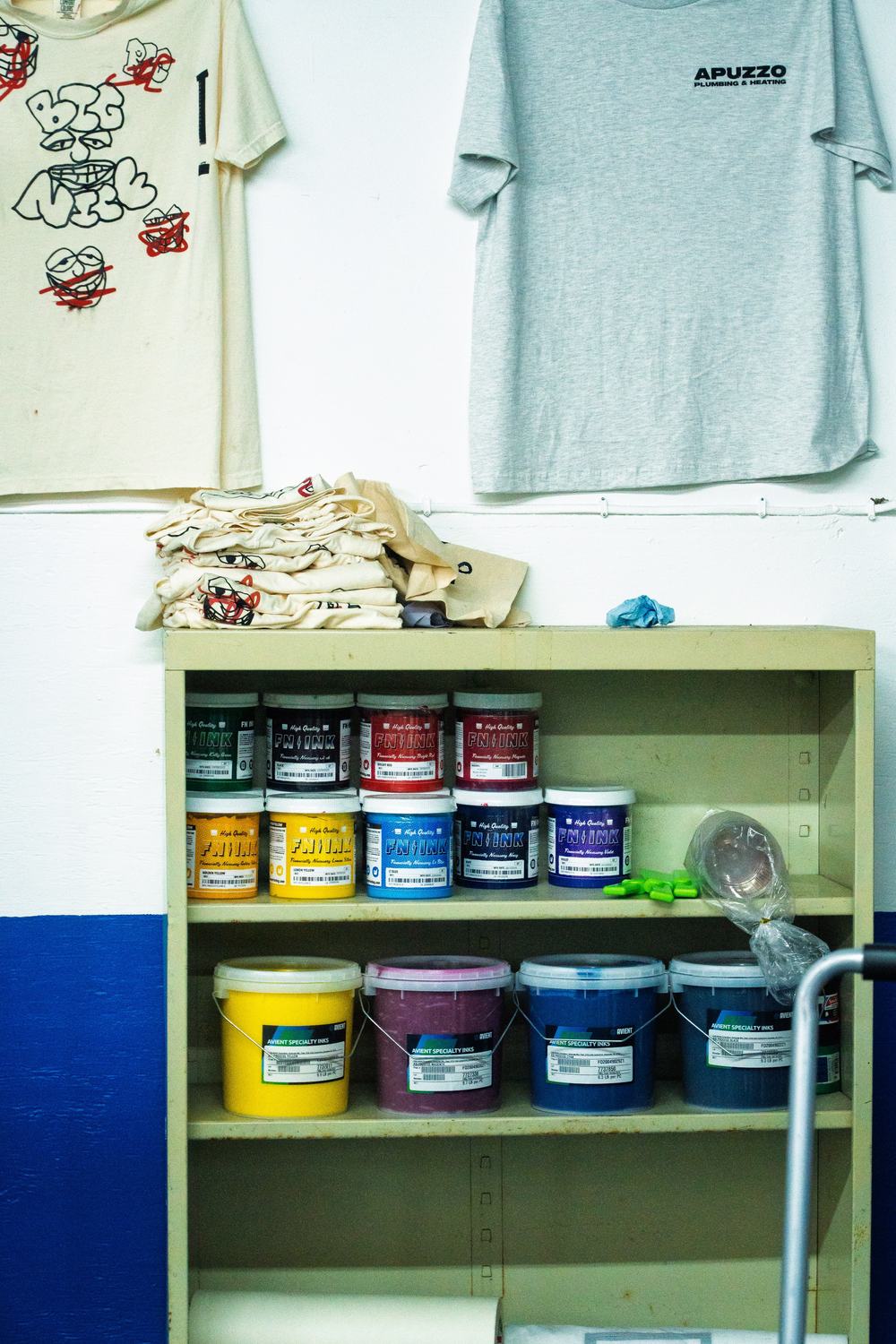Shelves of plastisol ink in quart jars and gallon buckets, under a Big Nice Studio tote print and an Apuzzo Plumbing & Heating work shirt.